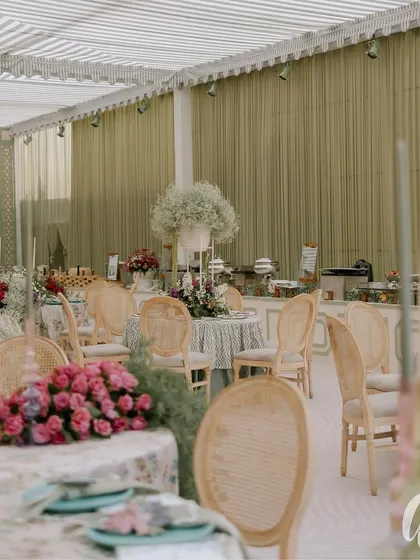 A wide view of the luncheon space, showing the striped marquee ceiling and sage green curtained walls. The layout provides a mix of round tables for an intimate and communal dining experience.