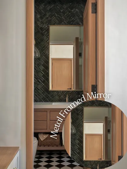 A metal-framed mirror adds a touch of industrial chic to this bathroom. The reflection shows the warm wooden door, creating a beautiful contrast with the dark herringbone tiles and checkerboard floor.