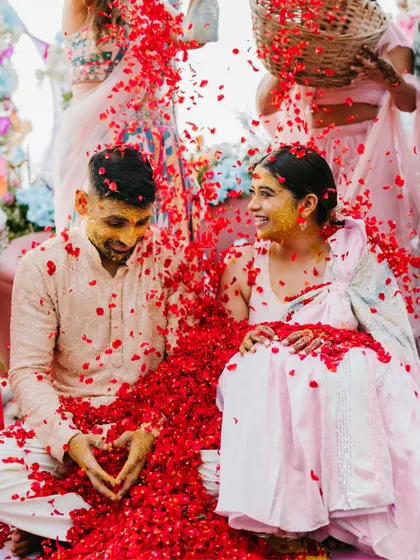 A joyful Haldi moment where the couple is showered with red rose petals, with the groom making a heart shape with his hands.