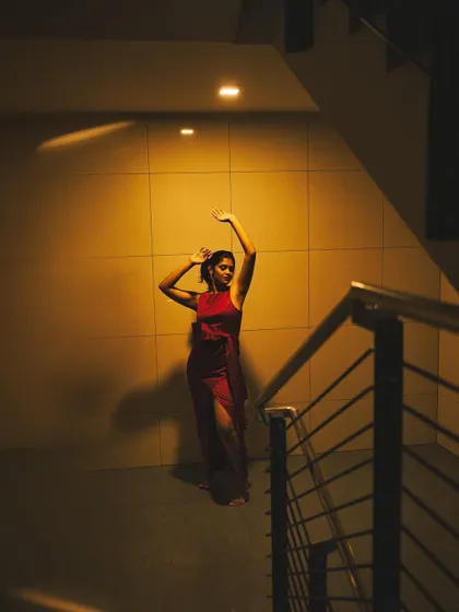 A dramatic, full-body portrait of a woman in a red dress posing under a single spotlight in a stairwell. The strong contrast between light and shadow creates a theatrical and powerful image.