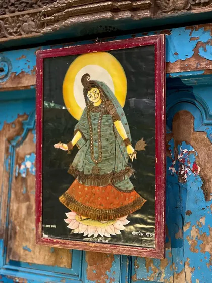 This cloth oleograph, featuring a deity against a full moon, is framed in a deep red that contrasts beautifully with the distressed blue door. Each piece is a carefully curated blend of color, texture, and tradition.