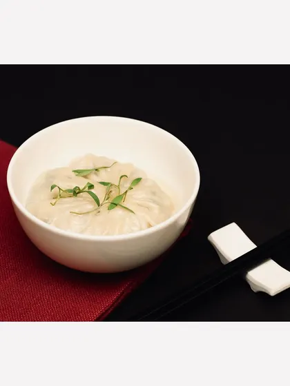 A simple and elegant presentation of steamed dumplings in a white ceramic bowl. The minimal styling with a red napkin and chopsticks on a black background gives it a clean, contemporary Asian dining feel.