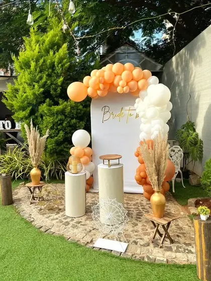 A simple yet beautiful rustic bridal shower backdrop. The design uses a white arch panel, an organic arrangement of orange and white balloons, and dried pampas grass for an earthy, natural feel.