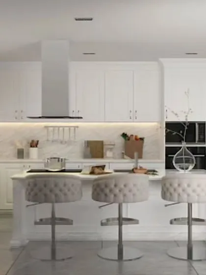 An elegant, all-white kitchen featuring a central island with comfortable, upholstered bar stools. Under-cabinet lighting and a marble backsplash add a touch of luxury and warmth to the classic design.