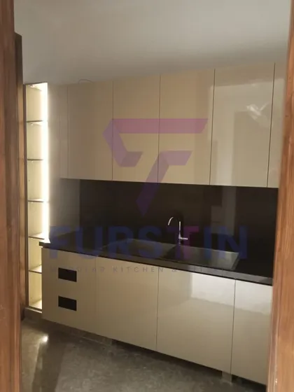 This part of the Bharatpur kitchen project features a dedicated wet area with a sleek, under-mount sink. The high-gloss beige cabinets are paired with a dark backsplash, creating a striking contrast, while the open, lit-up shelving unit adds a decorative touch.