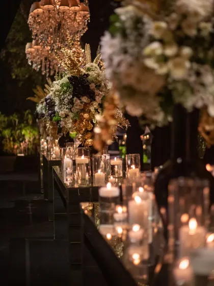 A long, mirrored table reflects an endless path of candlelight, flanked by towering arrangements of black, white, and gold florals. This design creates a powerful sense of depth and luxury for a sophisticated cocktail evening.