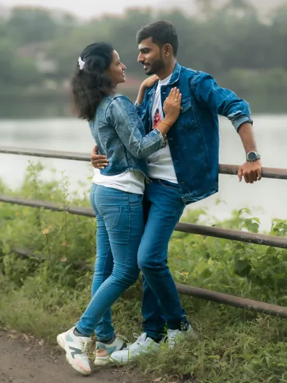 A casual and intimate pre-wedding photo of a couple in matching denim jackets, embracing by a lakeside railing.
