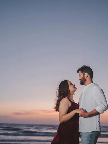 An intimate moment between a couple, holding hands and looking at each other against the backdrop of a soft sunset over the sea.
