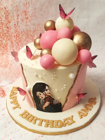 We create a personalized and heartfelt birthday cake for "Amma" with this beautiful design. A fault line reveals a cherished photo, while the cake is decorated with pink and gold spheres and delicate butterflies, blending personal touches with artistic flair.