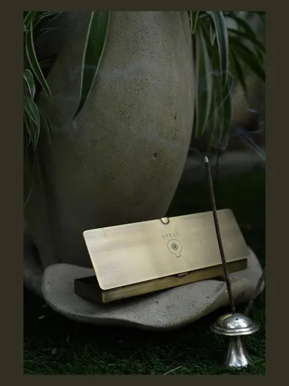 The gentle curl of smoke from an incense stick creates a serene mood. This shot for Byaas Home focuses on their elegant brass incense case, styled in a tranquil green setting to evoke a sense of calm.