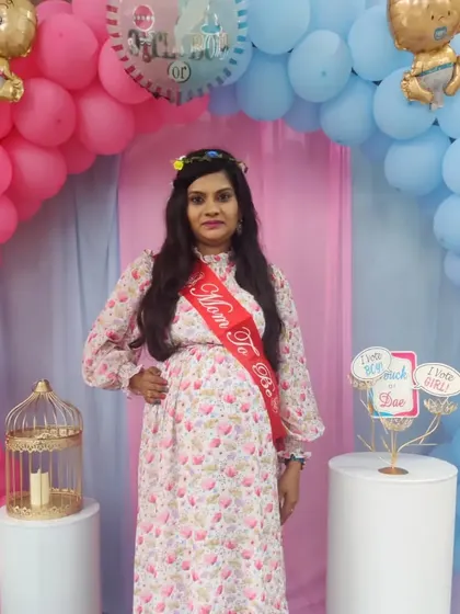 A happy "Mom To Be" posing in front of her baby shower decor. The pink and blue balloon arch and props create a festive and joyful atmosphere for the celebration.