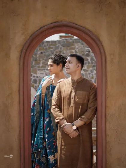 Framed within a rustic doorway, this portrait uses natural light to highlight the rich textures of the couple's ethnic outfits and the historic stone walls.