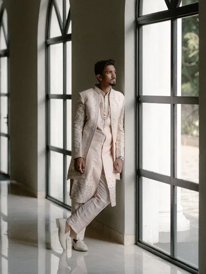 A groom leans against a row of windows, looking out thoughtfully. The natural light and strong lines of the architecture create a powerful and contemplative portrait.