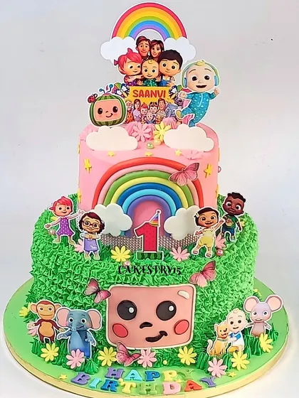 A vibrant two-tier Cocomelon cake for a first birthday. The design features a pink top tier and a green bottom tier with a grassy texture, decorated with numerous characters.