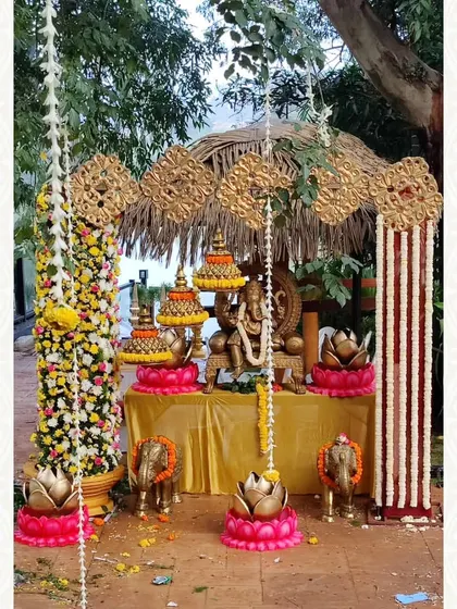 An outdoor Ganesha pooja setup under a rustic canopy. The idol is surrounded by lotus-themed decor, floral pillars, and small elephant figures, creating a serene and natural worship space.