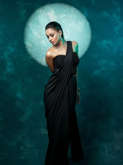 A full-length portrait set against a cool, moon-like backdrop. The model's pose and the dark, elegant saree create a feeling of celestial glamour.