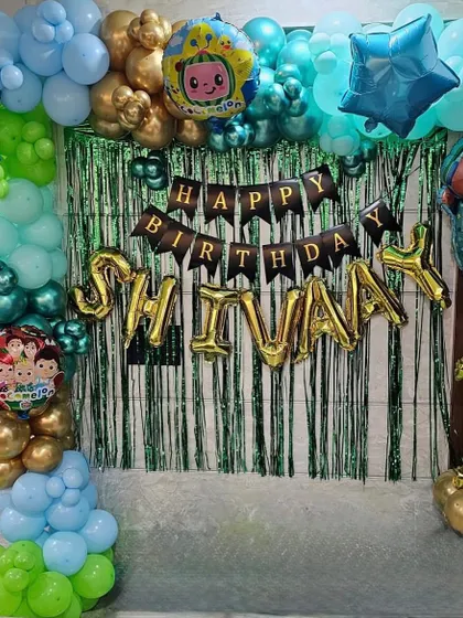 A Cocomelon themed backdrop for Shivay's first birthday. The decoration features a balloon arch over a green shimmer curtain, with the child's name in gold foil balloons.