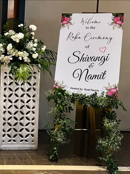 A personalized welcome sign for Shivangi and Namit's Roka ceremony. The elegant sign is placed on an easel and adorned with a garland of fresh greenery and flowers.