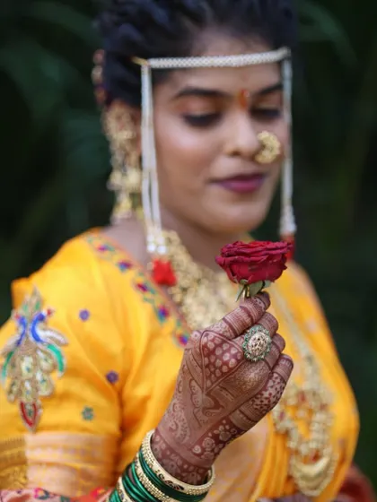 A close-up of the bride's hand, holding a single red rose. The dark henna stain provides a beautiful backdrop for her wedding jewelry.