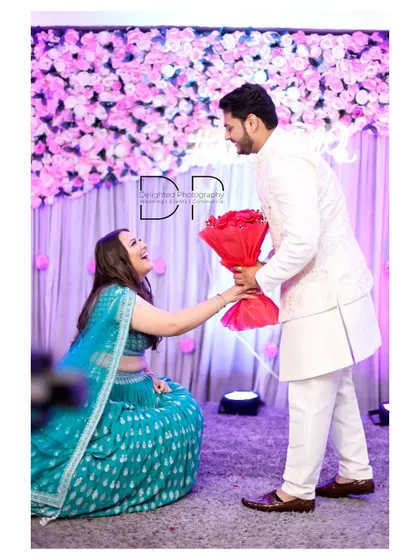 A modern twist on a proposal, with the bride-to-be on one knee presenting a bouquet of red roses to her partner during their engagement ceremony.