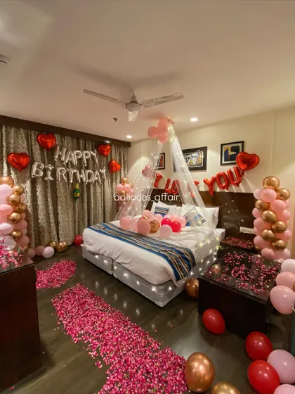 A full room view showing the balance of balloon columns, floor petals, and bed decorations in this rose gold themed birthday surprise.