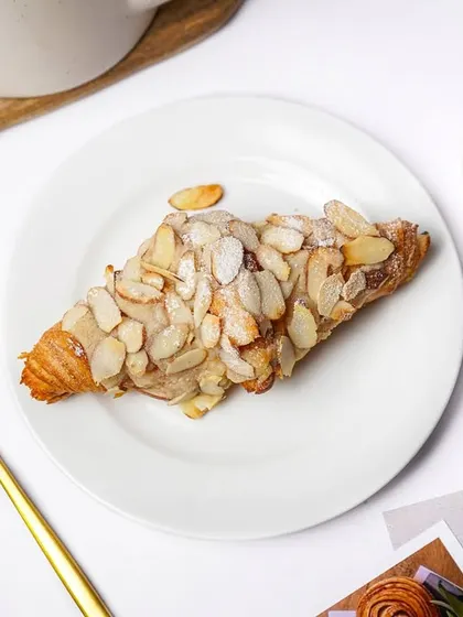 A flaky almond croissant, generously topped with sliced almonds, presented on a white plate with golden cutlery. This is an elegant shot for a high-end bakery or cafe.