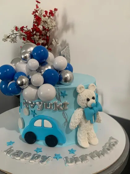 A cute first birthday cake with a teddy bear and car theme. The blue cake is decorated with silver, blue, and white chocolate balls for a festive look.