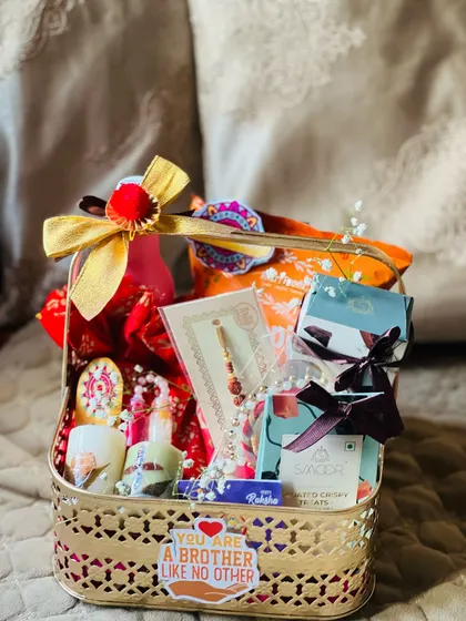 A golden metal basket filled with Rakhi goodies, including a rakhi, chocolates, and a "You are a brother like no other" sticker.
