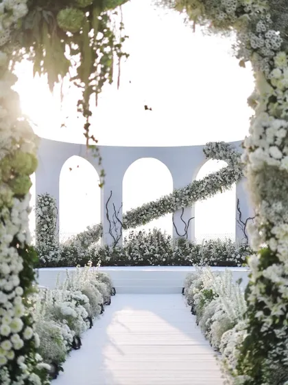 The wedding aisle, lined with a garden of white flowers, leads to the magnificent architectural mandap. This view, framed by a floral arch, creates a sense of anticipation and grandeur.
