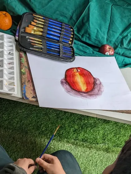 A student's perspective while working on a watercolor painting of an apple. This shot shows the reference object, the initial sketch, and the painting in progress.