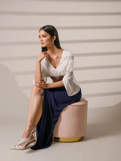 A full-length portrait of a model seated on a pink ottoman, wearing an elegant outfit. The clean studio lighting and shadows create a sophisticated, high-fashion look.