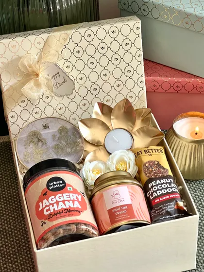 An elegant cream-colored Diwali box featuring a Ganesh and Laxmi resin stand, jaggery chana, and a lotus-shaped tealight holder.