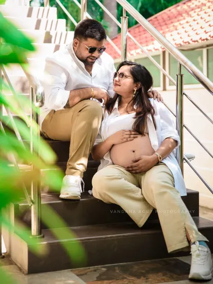 A relaxed and cool shot of a couple sitting on stairs outdoors. The mother-to-be is wearing a chic, casual outfit that proudly displays her baby bump.