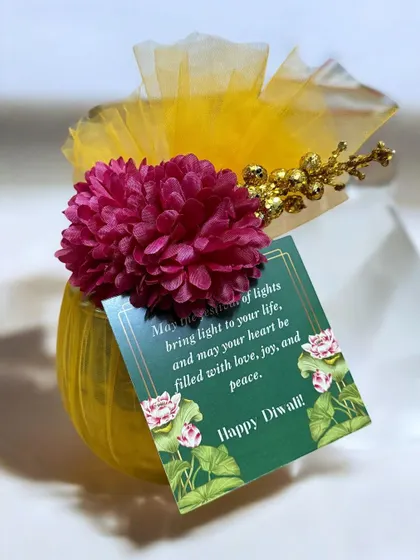 A close-up of the festive packaging for a small Diwali favor. A simple jar is elevated with bright yellow netting, a flower, and a custom "Happy Diwali" tag.