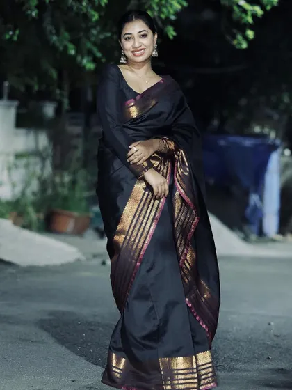 An outdoor, evening shot of a model in a black saree with a pink and gold border. The natural setting gives it a candid, lifestyle feel.