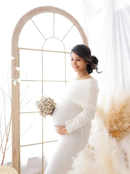 A light and airy portrait by the window. The mom-to-be holds a bouquet of dried flowers, her white lace dress glowing in the natural light.