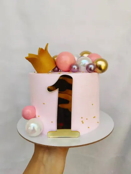 A pretty pink cake for a first birthday. The design is simple and elegant, with a gold crown, a large number one, and decorative spheres.