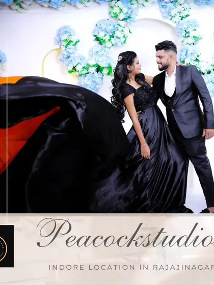 A romantic pre-wedding studio shot. The couple is dressed in matching black formal wear, with the woman's trail gown creating a dramatic effect against a floral arch.
