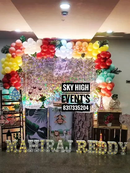 A wild birthday setup for Mahiraj Reddy, featuring a shimmering silver backdrop framed by a colorful balloon arch and various jungle animal props like an elephant and a leopard.