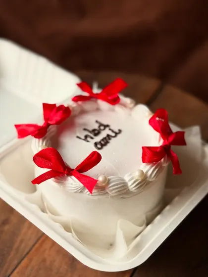 A classic white birthday bento cake for "Aai" (mom). It's decorated with delicate piping and four pretty red satin bows, giving it a timeless, gift-wrapped look.