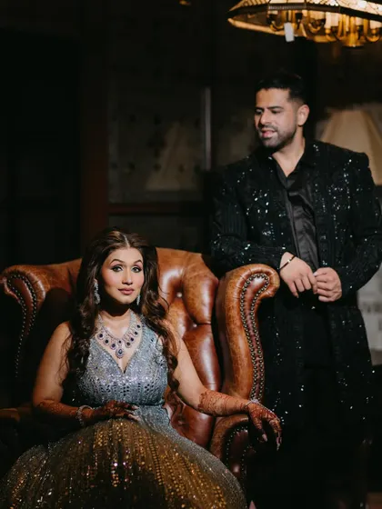 A stylish portrait of the couple in a classic room, with the bride seated in a leather armchair and the groom standing beside her, exuding elegance.