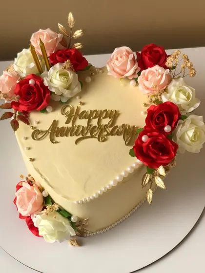 A romantic heart-shaped anniversary cake, framed with red, pink, and white roses and a golden topper.
