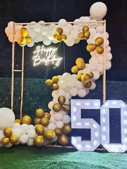 A stylish outdoor setup for a 50th birthday or anniversary. The gold and white balloon garland on a metal frame is paired with large marquee numbers.