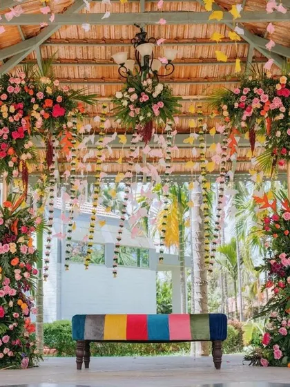 A full view of the stunning engagement stage. The combination of a multi-colored bench, lush floral pillars, and a canopy of hanging paper flowers creates a whimsical yet elegant atmosphere.