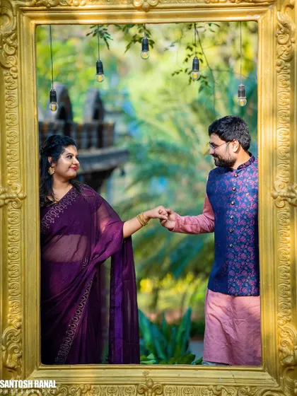 A creative pre-wedding shot using a large golden frame to capture the couple in a garden setting.