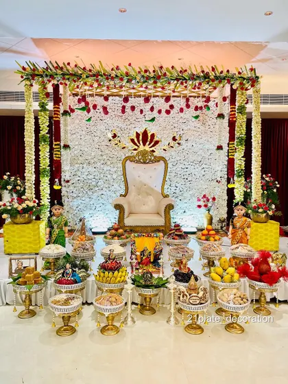 An elegant stage setup with a white floral wall and a lotus motif above the throne. The decor is framed by traditional pillars and includes a full display of my signature plates.