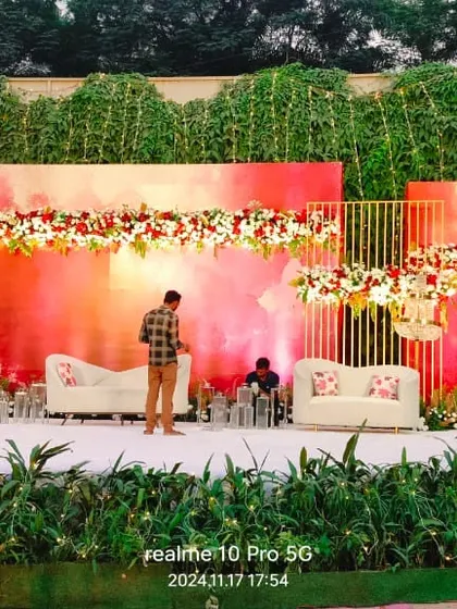 A modern and artistic reception stage setup at The Acres Club. The watercolor-style backdrop in shades of pink and red, paired with clean white sofas, offers a contemporary and vibrant look.