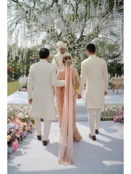 A walk to remember. The bride, escorted by her brothers, makes her way down a pristine white aisle lined with soft pastel flowers, heading towards a beautiful garden mandap for a serene and elegant farmhouse wedding ceremony.