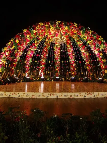 A closer look at the magnificent floral dome mandap at night. The internal lighting and surrounding spotlights make the vibrant flowers glow, transforming the space into a magical, illuminated sanctuary for the couple.