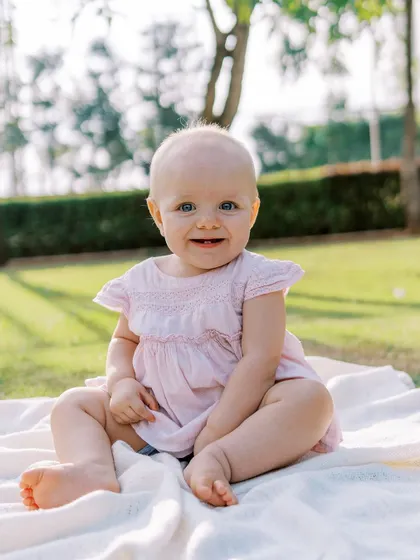 A happy baby girl with a sweet, toothy smile, sitting on a blanket outdoors. Her joy is absolutely radiant.